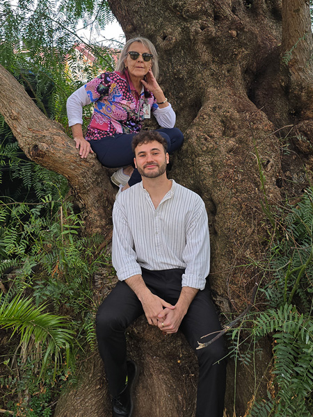 A woman sits on a tree branch while a man sits below her at the tree’s base, surrounded by green foliage.