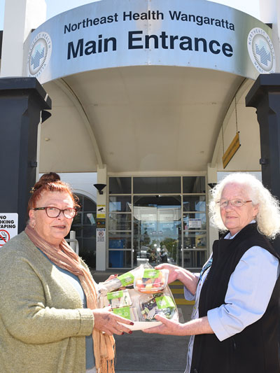 Two older ladies holding up a selection of food and stand outside NHW main entrance.