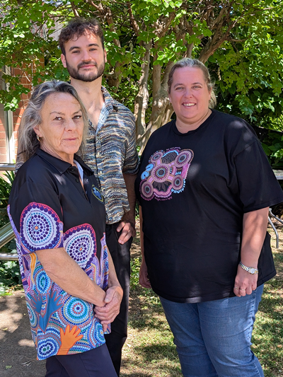Three people stand outdoors under a tree, smiling at the camera. They wear shirts with an aboriginal design. These people are from our Aboriginal Health team.