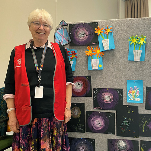 Rosie is stood in front of a picture board full of artwork created by the residents at Illoura Aged Care