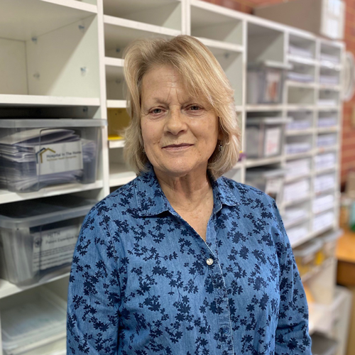 NHW Volunteer Rhonda Taylor is wearing a dark blue flowered shirt and stood in front of shelving staked with information sheets and booklets. 