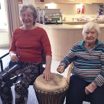 Two elderly females sat down with a djembe drum between them while listen to local drumming group